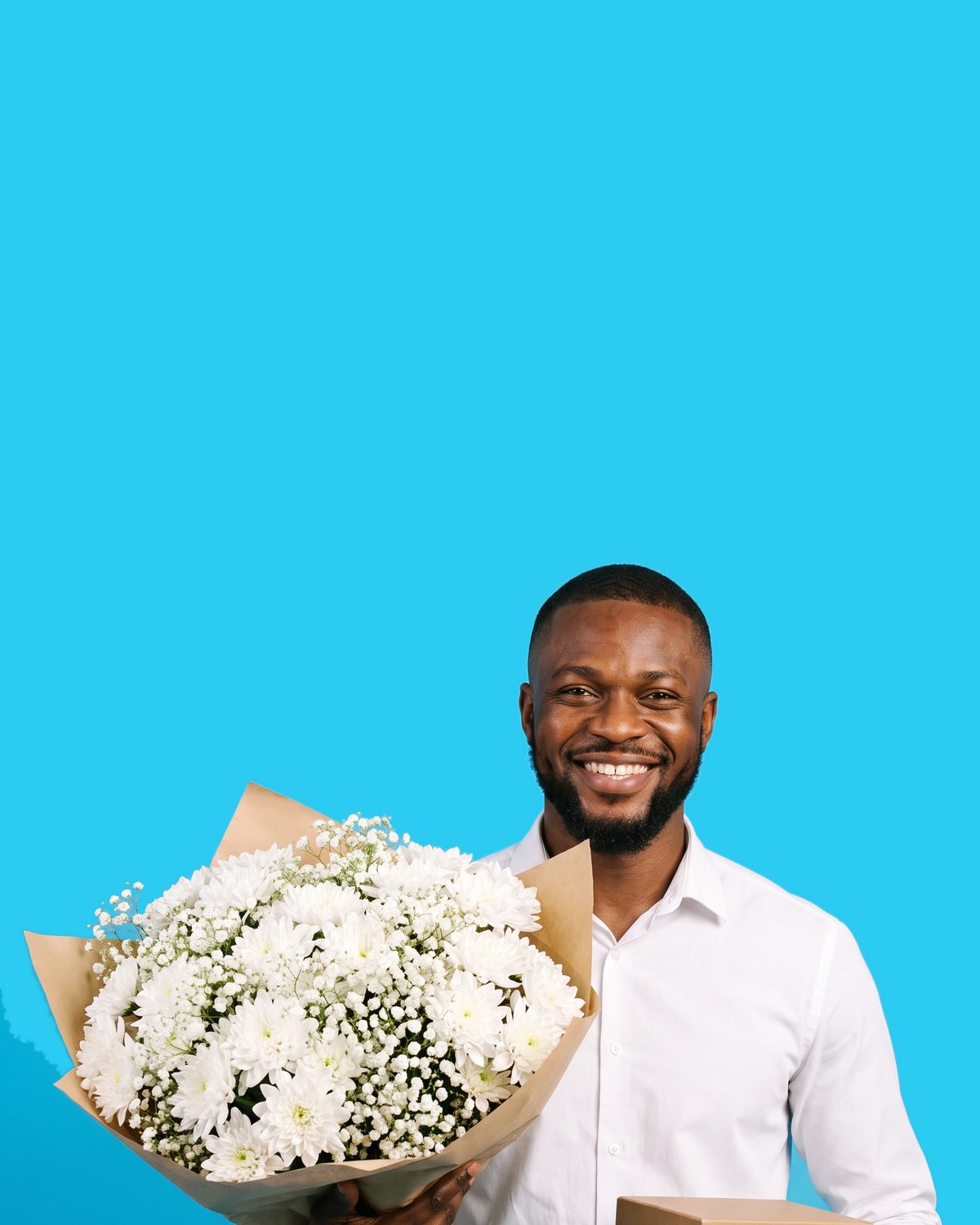 A man grinning, holding a wrapped gift box in one hand and a small bunch of white chrysanthemums in the other