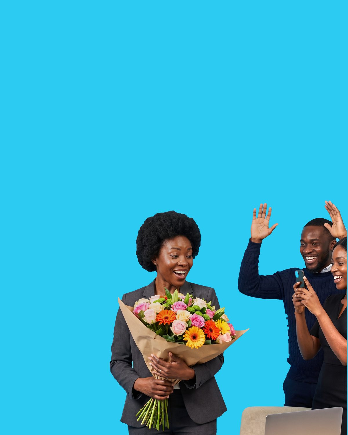 A woman in a grey suit laughing as she holds a kraft-wrapped bouquet of pink and cream roses with orange and yellow gerberas, two colleagues cheering behind her with their arms raised, one of them filming on a phone