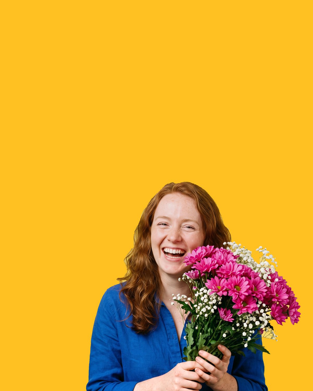 A woman with long red hair in a blue shirt, laughing, holding a bouquet of hot-pink chrysanthemums and gypsophila