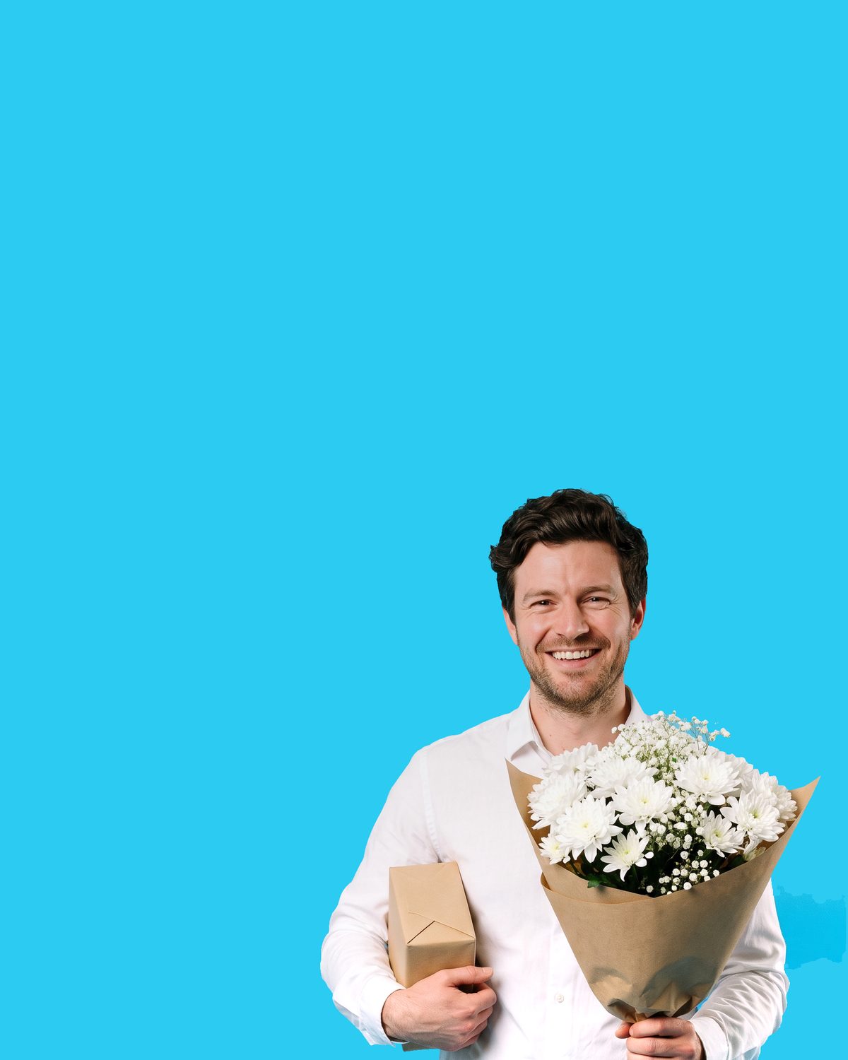 A dark-haired bearded man in a pale shirt, smiling, holding a wrapped bouquet of white daisies and a small kraft gift box