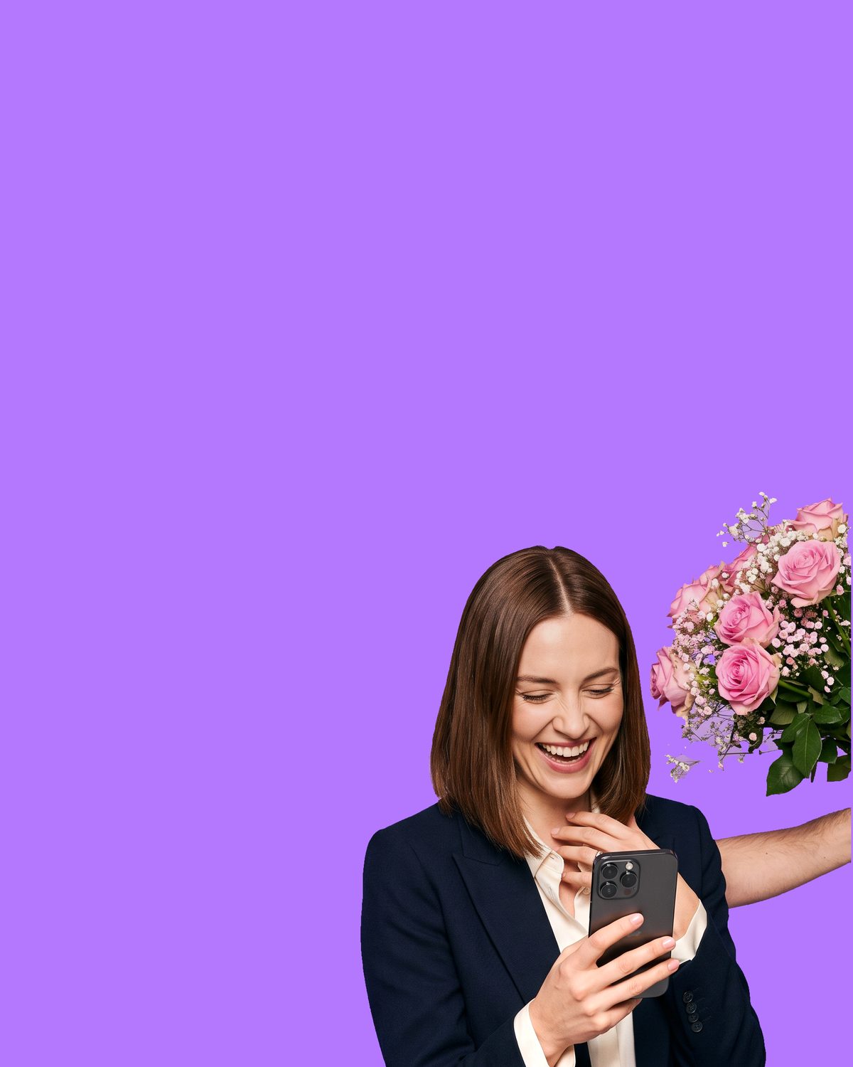 A woman with a shoulder-length brown bob laughing at the phone in her hand, her other hand at her collar, as a bouquet of pink roses and gypsophila is held out to her from off-frame
