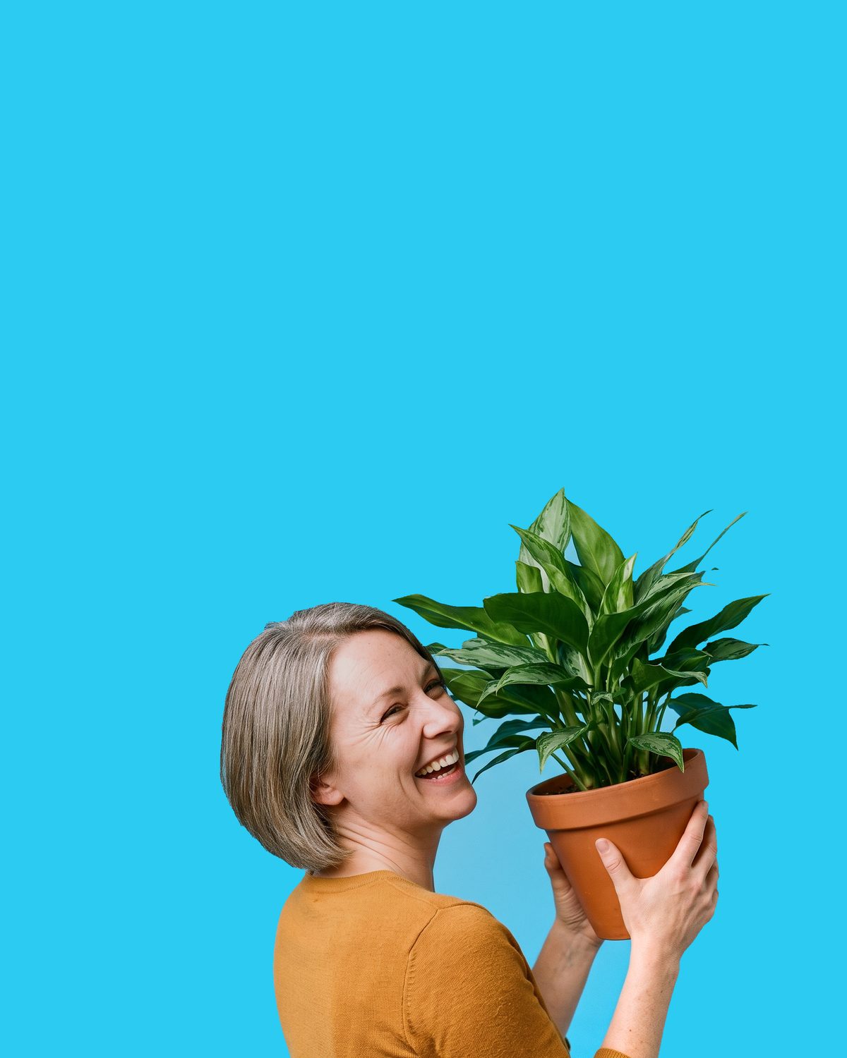 An older woman with grey bobbed hair in a mustard top, eyes closed and smiling, holding a potted peace lily in a terracotta pot