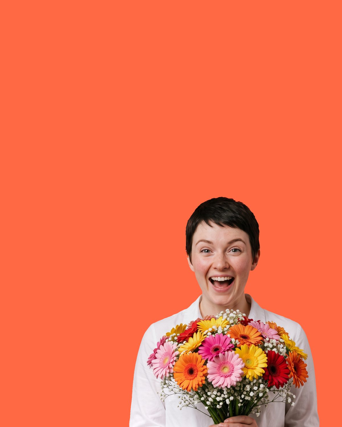 A woman with short dark hair in a white shirt, delighted, holding a bright bouquet of pink, orange and yellow gerberas