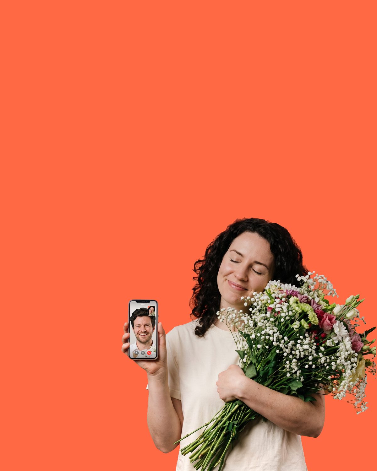 A woman with dark curly hair, eyes closed, hugging a bouquet of white flowers and gypsophila in one arm and holding up her phone in the other, a man smiling back from the video call on its screen
