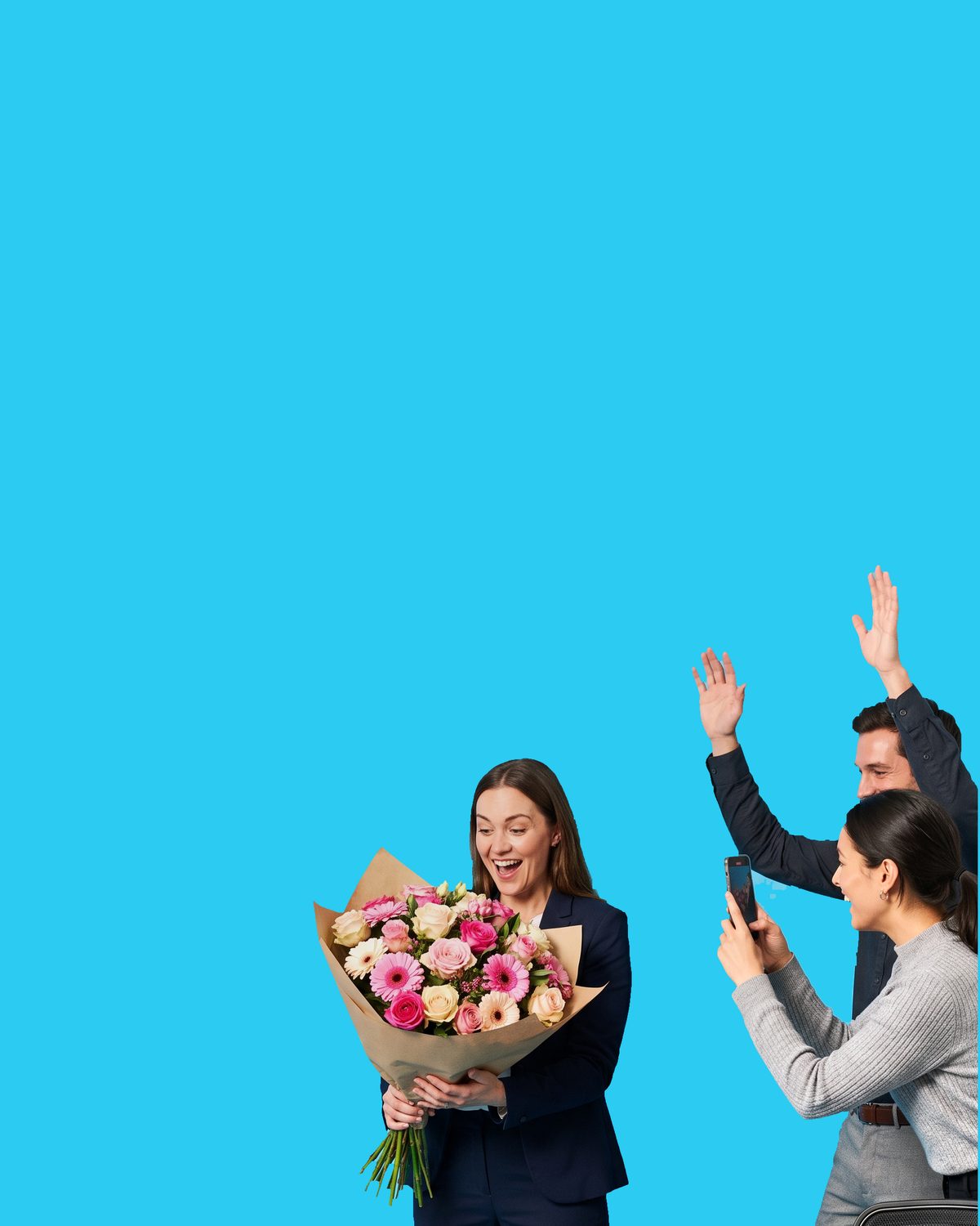 A woman in a navy suit laughing down at a kraft-wrapped bouquet of pink and cream roses and gerberas in her arms, two colleagues behind her with their hands in the air, one of them filming on a phone
