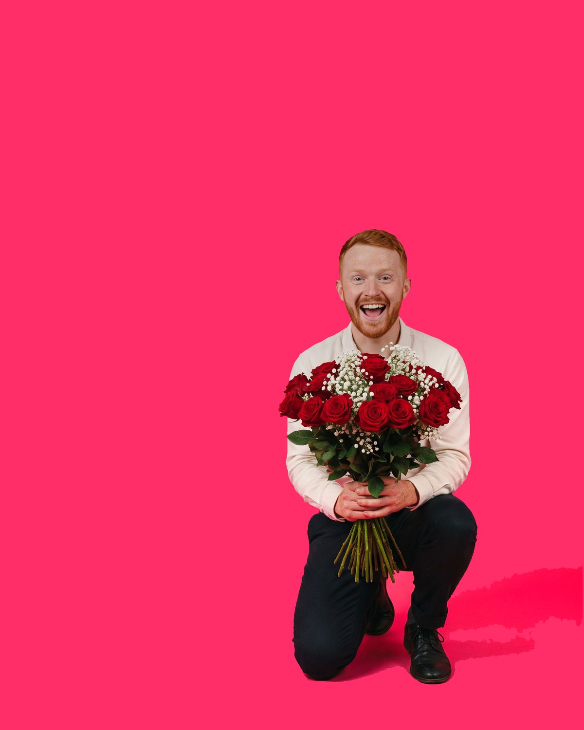 A red-haired bearded man in a white shirt down on one knee, laughing, holding a large bouquet of red roses